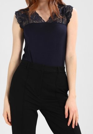 Top sin mangas de color azul marino con bordes de encaje en los hombros, combinado con pantalones negros de cintura alta con pliegues frontales.