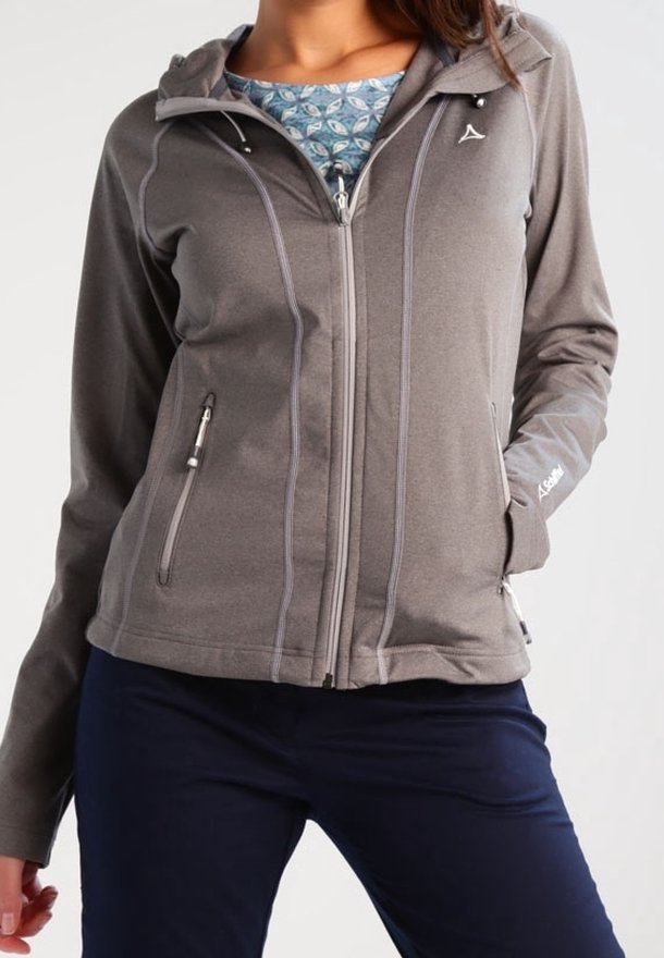 Femme portant une veste grise zippée avec des poches à fermeture éclair et une chemise bleue à motifs en dessous, associée à un pantalon bleu marine.