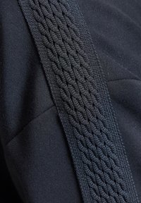 Gros plan d'une sangle en tissu tressé noir posée sur un tissu sombre texturé avec des coutures et des lignes de couture visibles.