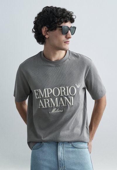 Gray short-sleeve T-shirt with textured fabric and "Emporio Armani Milano" logo in cream. Paired with light blue denim jeans and sunglasses.