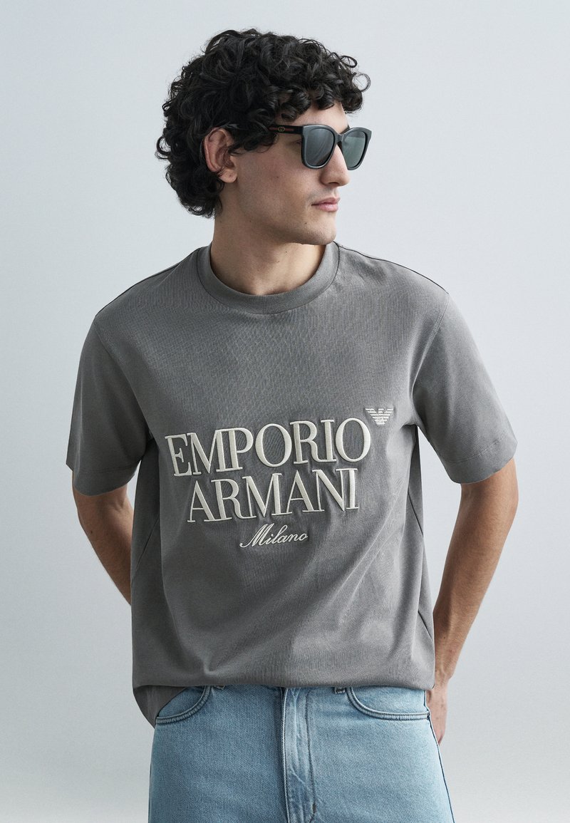 T-shirt cinza de manga curta com tecido texturizado e logótipo "Emporio Armani Milano" em creme. Combinado com jeans de ganga azul claro e óculos de sol.