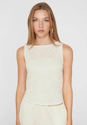 Joven mujer con cabello rubio liso lleva una blusa blanca sin mangas y pantalones blancos a juego, posando frente a un fondo claro y simple.