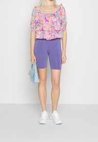 Blouse à manches courtes à motif floral rose et violet, design à couches superposées ; associée à un short de vélo violet. Tient un sac à main bleu clair. Baskets blanches.