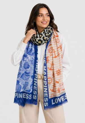 Frau trägt weißes Hemd und beige Hose, kombiniert mit zwei Schals: einer mit Leopardenmuster, einer mit blau-orangefarbenem nautischem Muster mit den Worten „Happiness“ und „Loves“.