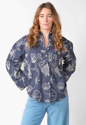 Femme aux longs cheveux bouclés portant une chemise bleu marine à boutons avec des motifs blancs de vie marine et un jean bleu clair, debout les mains sur les hanches.