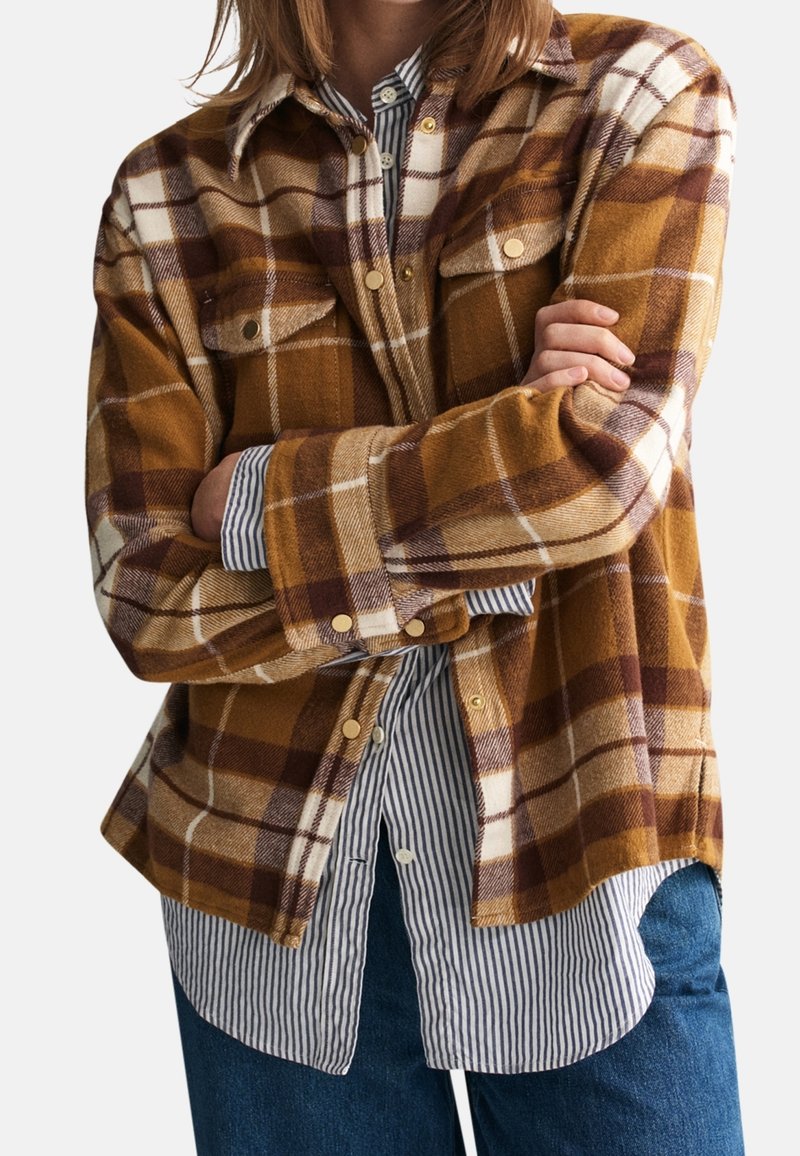 Camisa de franela a cuadros marrón y amarilla con dos bolsillos en el pecho, botones dorados y una camisa de rayas azules y blancas superpuesta debajo.