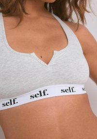 Svetlo šedý športový bralet s výstrihom do V a bielym pásom s nápisom "self." v čiernej farbe. Hladká textúra a minimalistický dizajn.