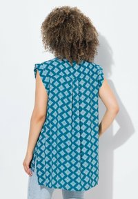Türkise ärmellose Bluse mit weißem Diamantmuster, ausgestattet mit einem Rückenschlitz und seitlichen Schlitzen für mehr Bewegungsfreiheit.