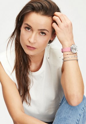 Silberne Uhr mit rundem Zifferblatt und pinkem geflochtenem Lederarmband. Modell trägt ein weißes ärmelloses Shirt und Jeans aus Denim, mit silbernen Accessoires.