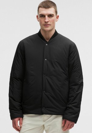 Uomo che indossa una giacca imbottita nera con colletto alla coreana e pantaloni bianco sporco, in piedi davanti a uno sfondo chiaro e uniforme.