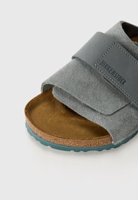 Sandale en suède gris avec sangle ajustable, semelle en liège et semelle extérieure en caoutchouc teal. Présente un dessus texturé et une surface intérieure lisse.