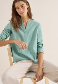 Jeune femme aux longs cheveux bruns portant un chemisier vert clair texturé et un pantalon beige, assise de manière détendue sur une chaise en bois devant un fond uni.