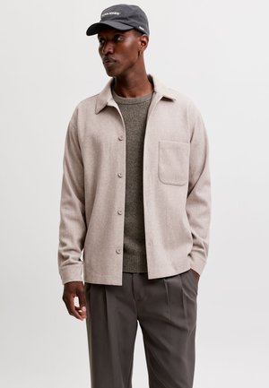 Homme portant une casquette grise, une veste beige boutonnée sur un pull marron et un pantalon gris foncé, debout avec une main dans la poche.