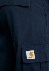 Granatowa koszula robocza wykonana z teksturowanej tkaniny, z kwadratową kieszenią, na której znajduje się białe logo Carhartt oraz pomarańczowy akcent.
