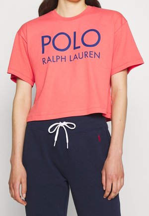 Person trägt ein korallenfarbenes, kurzes T-Shirt mit dem Schriftzug "POLO RALPH LAUREN" und dunkelblaue Jogginghosen mit weißem Kordelzug und kleinem rotem Logo.