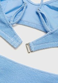 Modra teksturirana kopalna obleka, ki vključuje bralette z nastavljivimi naramnicami in zapenjanjem, dopolnjena z usklajenimi hlačkami. Mehka, plišasta tkanina.