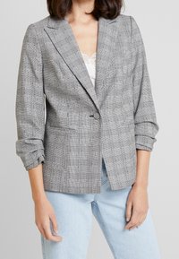 Svart- och vitt rutig blazer med en knapp, notched lapels och uppvikta ärmar, bärs över en topp med spetsdetaljer och ljusa jeans.