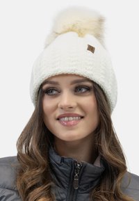 Bonnet en tricot blanc avec un pompon duveteux sur le dessus, en matière texturée, et une petite étiquette en cuir marron sur le côté.