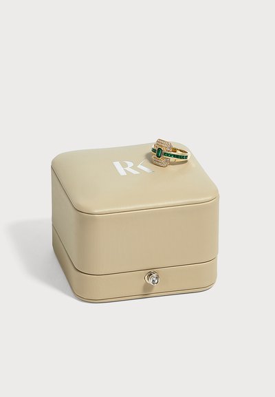 Bague en or avec des pierres précieuses vertes et transparentes reposant sur une boîte à bijoux beige avec un logo RK blanc et une petite poignée de tiroir.