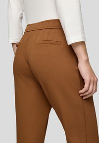 Braune strukturierte Hose mit taillierter Passform, die einen glatten Bund und Gesäßtaschen aufweist. Das Material wirkt weich und flexibel.