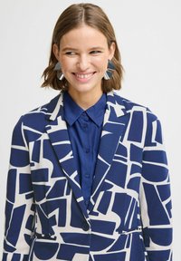 Blazer avec un motif géométrique bleu et blanc, doté d'un col cranté et d'une fermeture à un bouton, associé à une chemise bleue unie.