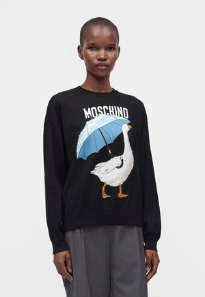 Γυναίκα που φοράει μαύρο πουλόβερ Moschino με λευκή χήνα που κρατά ένα μπλε ομπρέλα και γκρι παντελόνι με φαρδιά πόδια.