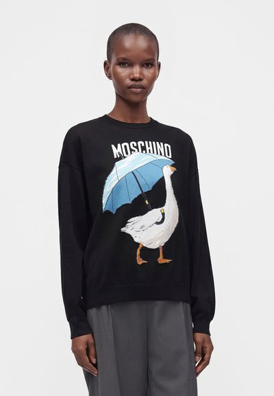 Γυναίκα που φοράει μαύρο πουλόβερ Moschino με λευκή χήνα που κρατά ένα μπλε ομπρέλα και γκρι παντελόνι με φαρδιά πόδια.