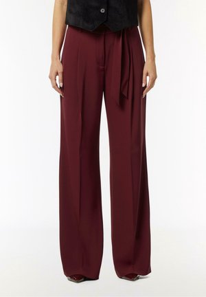 Frau trägt weinrote, hochtaillierte, weit geschnittene Hosen mit vorderen Falten und einem passenden Stoffgürtel, der locker an der Taille gebunden ist.