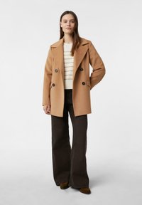 Dwurzędowy płaszcz z mieszanki wełny w kolorze wielbłądzim, z dużymi klapami i czarnymi guzikami. W zestawie z kremowym swetrem w paski i brązowymi, rozszerzanymi spodniami.