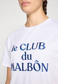 Weißes Baumwoll-T-Shirt mit blauem besticktem Text "le CLUB du MALBôn" auf der Vorderseite, rundem Ausschnitt und kurzen Ärmeln.