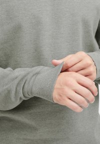 Suéter de punto gris con puños acanalados, mostrando manos ajustando la manga. Textura suave y diseño clásico de cuello redondo.