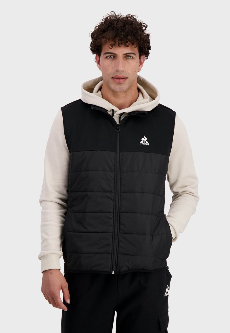Gilet matelassé noir avec fermeture éclair devant, col montant et logo sur la poitrine. Porté par-dessus un hoodie beige, avec une texture lisse et un design structuré.