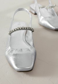 Hopeat metalliset slingback-kengät, joissa on neliön muotoinen kärki, koristeltu kirkkailla rhinestone-nauhoilla etuosassa ja matalalla korkokengällä.