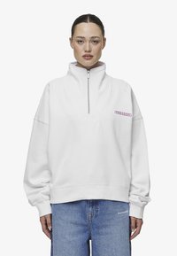 Sudadera blanca de forro polar con cuello alto, media cremallera y un diseño contorneado. Presenta un pequeño logo morado de 'PEGADOR' en el pecho.