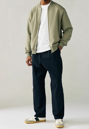 Homme portant une veste zippée vert olive, un t-shirt blanc, un pantalon foncé et des baskets blanches avec des accents noirs et beiges, debout avec une main dans la poche.