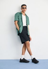 Camisa de manga curta verde claro, top branco de alças, calções pretos com logótipo, ténis pretos, meias brancas; de pé contra um fundo simples.