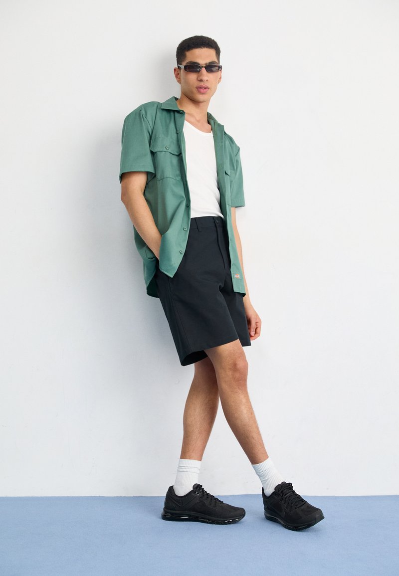 Camisa de manga corta verde claro, camiseta blanca de tirantes, pantalones cortos negros con logo, zapatillas negras, calcetines blancos; de pie contra un fondo liso.