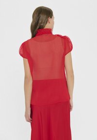 Czerwona, przezroczysta bluzka z falbaniastym kołnierzykiem i krótkimi bufiastymi rękawami. Tył ma gładkie wykończenie i subtelne przejścia faktury.