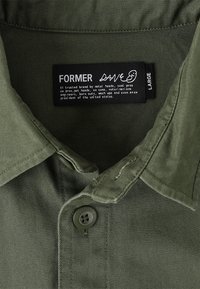 Olive green shirt with a folded collar, featuring a button closure. Tag indicates size "Large" and brand name "Former." Cotton fabric texture visible.