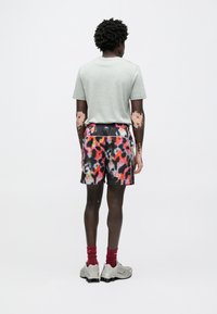 Γκρι κοντομάνικη μπλούζα, πολύχρωμα σορτς με μοτίβα σε ροζ, πορτοκαλί και μαύρο, κόκκινες κάλτσες, γκρι αθλητικά παπούτσια. Πλάγια όψη, απλό φόντο.