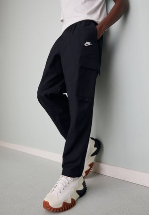 Persona vestida con pantalones cargo negros de Nike y zapatillas blancas con suelas gruesas y onduladas en marrón y negro, de pie contra una pared pálida.