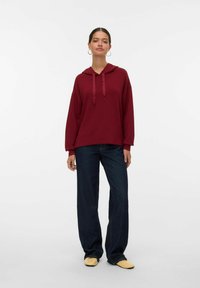 Sudadera burdeos con cuello de cordón, que presenta un corte relajado y mangas largas, combinada con jeans de pierna ancha azul oscuro y zapatos de ante amarillos.