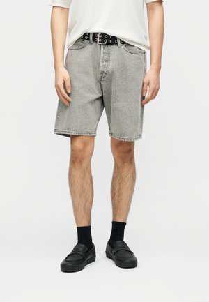Mann trägt hellgraue Jeansshorts, schwarzen Gürtel mit silbernen Ösen, schwarze Socken und schwarze Slipper, steht vor einem weißen Hintergrund.