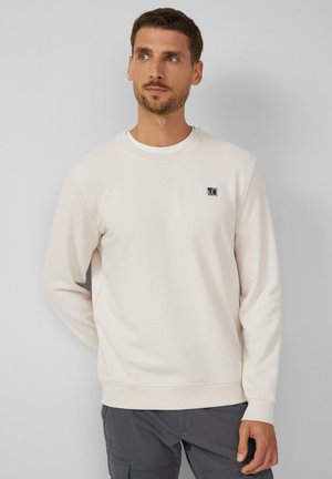 Sudadera beige claro con cuello redondo, mangas largas, puños acanalados y un pequeño parche de logo en el pecho. Tejido suave y texturizado.