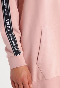 Rosa sweatshirt med kängurufickor, ribbade ärmslut och en kontrasterande svart- och vitlogo-stripe längs ärmen.