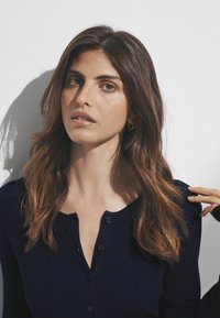 Top blu navy a coste con bottoni e scollatura a V. La modella ha i capelli lunghi e ondulati e posa su uno sfondo bianco uniforme.