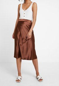 Bílé tílko s detailním knoflíkem, kombinované s hnědou saténovou sukní, která sahá do poloviny lýtek. Doplněno bílými skluznými sandály. Hladká textura.