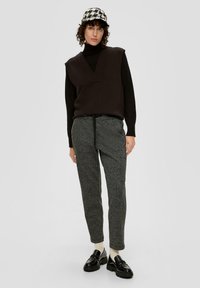 Zwarte mouwloze trui met V-hals, grijze gestructureerde broek en zwarte instappers. Het model draagt een geruite pet en crèmekleurige sokken.