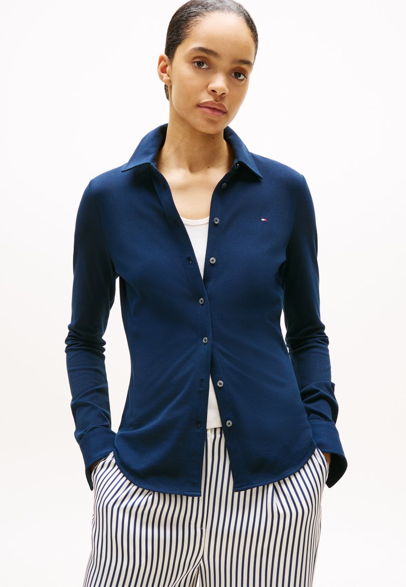 Mujer con una camisa azul oscuro abotonada sobre una camiseta blanca, con las manos en pantalones blancos con rayas azules verticales, de pie frente a un fondo liso.