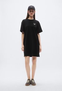 Čierne oversize tričkové šaty s krátkymi rukávmi a okrúhlym výstrihom, s bielym logom na hrudi a v kombinácii so sivými teniskami.